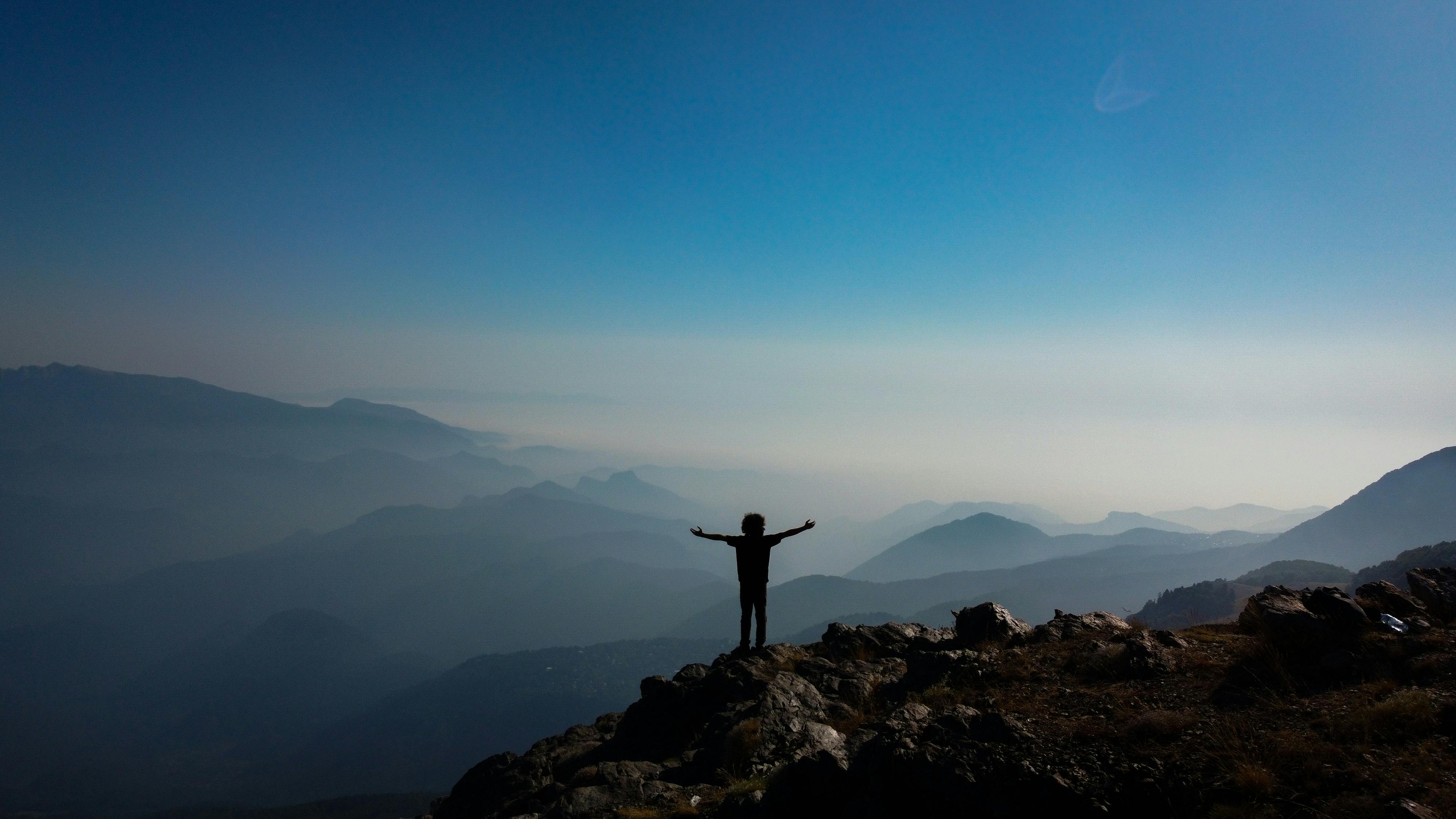 Person standing on a mountain summit with arms spread wide, overlooking misty blue ranges.
